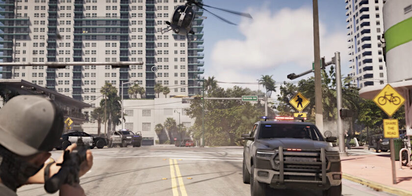 Screenshot aus dem Videospiel „GTA 6“ zeigt eine Verfolgungsjagd-Szene aus der Egoperspektive, bei der ein Charakter mit einem Sturmgewehr auf ein herannahendes Polizeifahrzeug zielt, während ein Polizeihubschrauber tief über die Straße fliegt. Im Hintergrund sind Hochhäuser einer Küstenstadt sowie weitere Polizeifahrzeuge zu erkennen.
